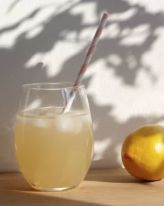 Un verre avec une citronnade est posée à gauche de l'image sur un table en bois. A sa droit un citron jaune. Le deux sur à l'ombre d'un arbre. A découvrir dans le Carnet des Explorations sensorielles du thé