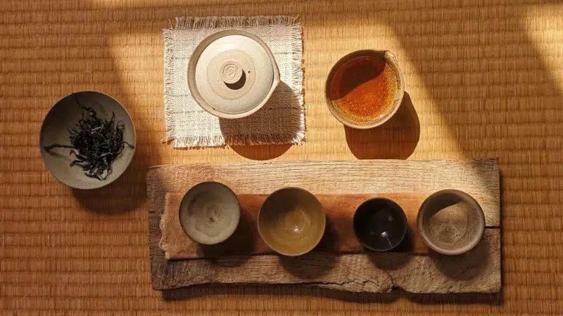 Photographie des céramiques de Manon Cluzeau sur un tatami. On peut appercevoir un gaïwan fermé, une verseuse, 4 tasses et un bol avec des feuilles de thés. Cette image est représentative de la vision de la feuille à la tasse, des techniques d'infusion traditionnelles et modernes.
