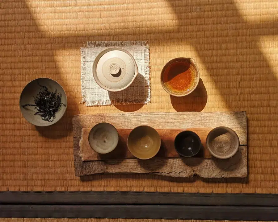 Photographie des céramiques de Manon Cluzeau sur un tatami. On peut appercevoir un gaïwan fermé, une verseuse, 4 tasses et un bol avec des feuilles de thés. Cette image est représentative de la vision de la feuille à la tasse, des techniques d'infusion traditionnelles et modernes.