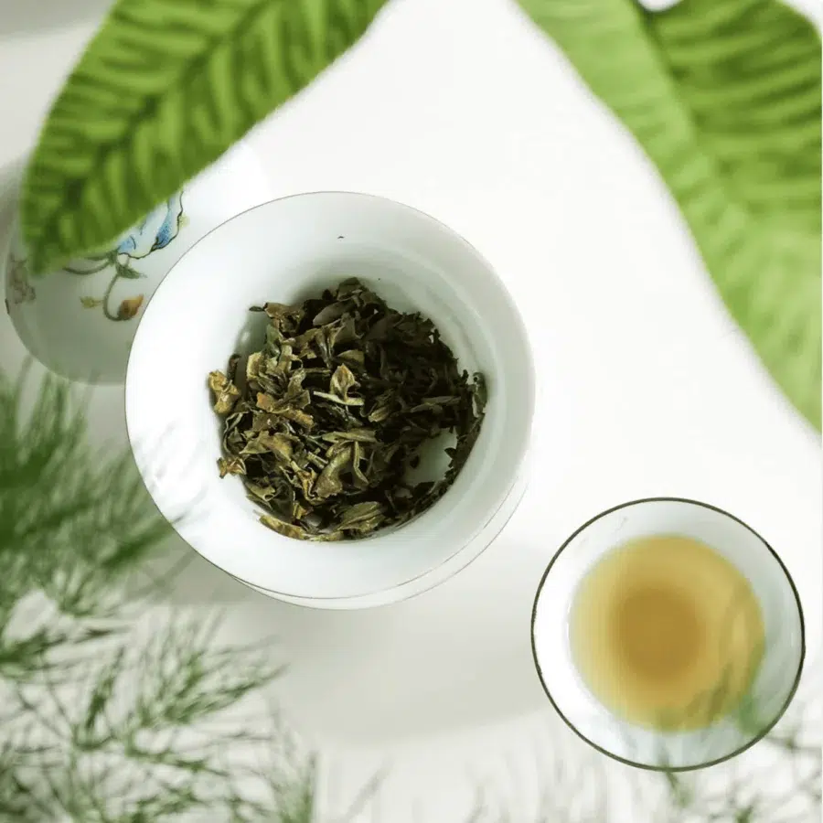 Gaïwan ( bol d'infusion chinois), le couvercle est ouvert. On aperçoit les feuilles de thé vert. En bas à sa droite, une tasse avec la liqueur jaune, celle du thé vert. Le deux objets sont dans un cadre entourés de 2 plantes. Atelier de thé sur mesure