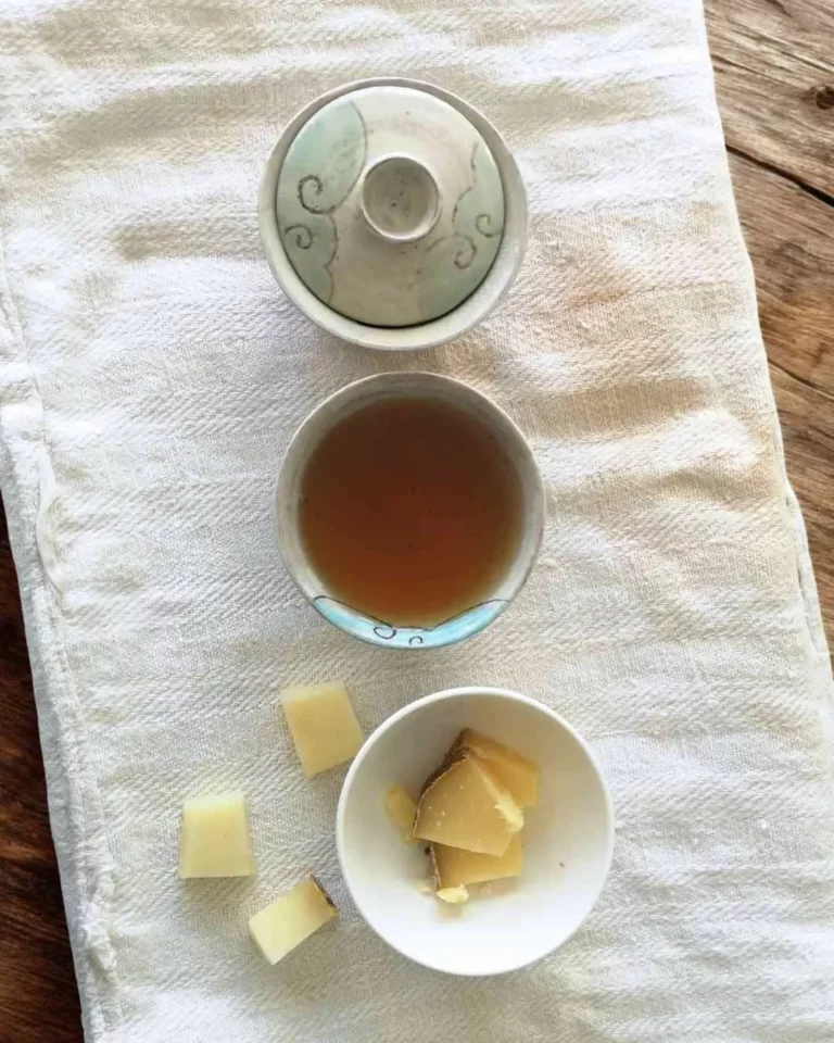 Atelier Fromages et Thés chez l'Affinez. Présentation sur une planche en bois et un tissu blanc, un gaïwan bleu gris avec en dessous une tasse de la même couleur, à l'intérieur une liqueur de thé noir. La partie inférieure un petit bol blanc avec des petits morceaux de fromages à pâte dure.