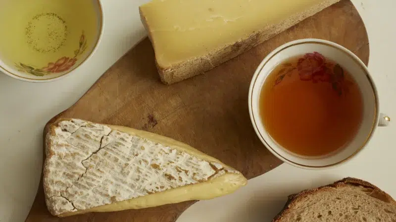 Photographie pour l'atelier Thés & Fromages chez Agatto. Deux fromages: un brie, un comté sur une planche en bois, avec une tranche de pain et deux tasses à thé en porcelaine.