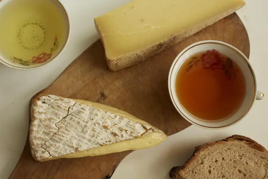 Photographie pour l'atelier Thés & Fromages chez Agatto. Deux fromages: un brie, un comté sur une planche en bois, avec une tranche de pain et deux tasses à thé en porcelaine.