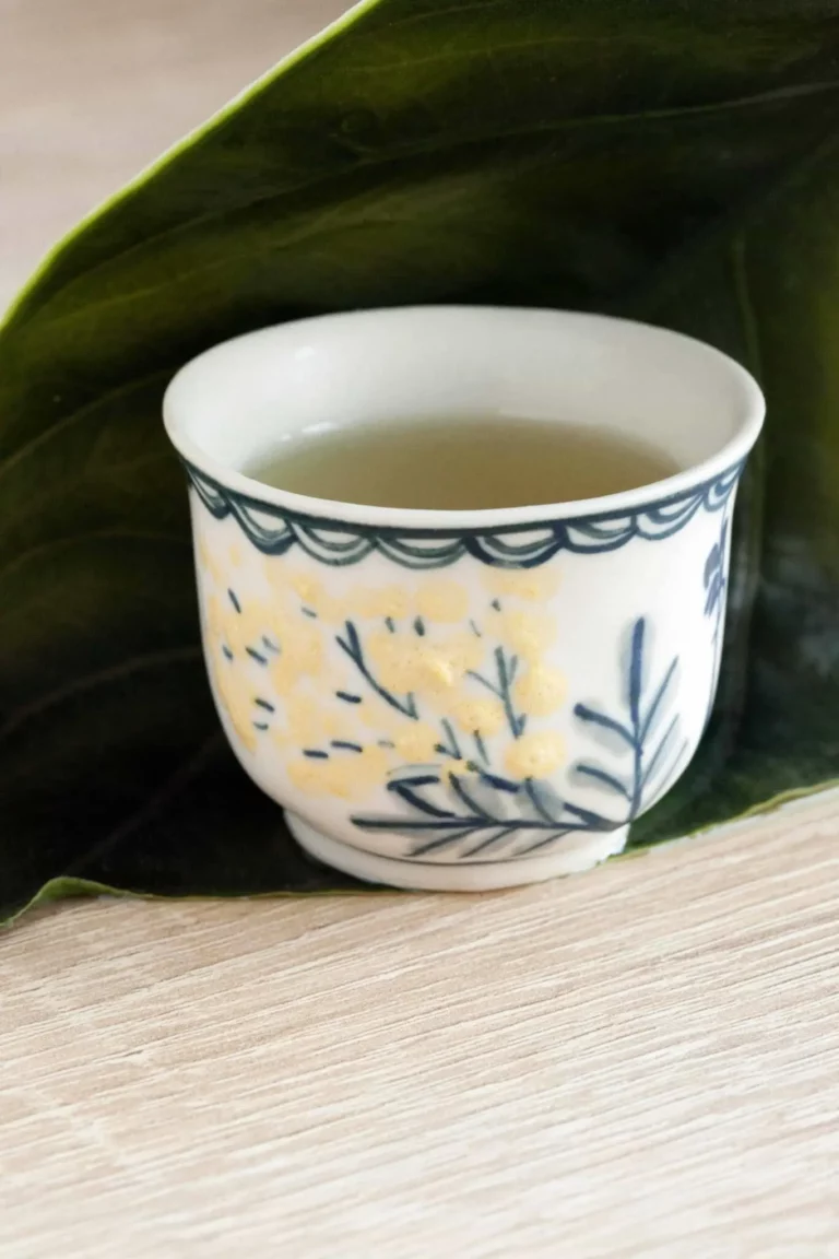 Photographie d'une tasse à thé de petit format avec du mimosa peint dessus. La tasse est posée sur une planche en bois et une grande feuille verte. 'Offres thé personnalisées: le thé à votre image'